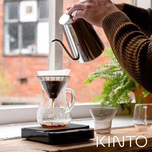 一只手正從不鏽鋼手沖咖啡壺中，將熱水注入玻璃濾杯，濾杯內有濾紙及咖啡粉。下方是透明玻璃咖啡分享壺，置於黑色電子秤上。背景是窗戶與綠色植物。