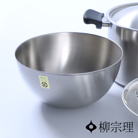 一個銀色不鏽鋼調理盆，盆壁光滑，邊緣有細微反光。盆側貼有一張黃色貼紙，上面印有黑色圖案與文字。盆後方可見一個同樣材質的鍋子，鍋蓋上有黑色把手。