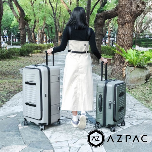 一位留著中長黑髮的女性背對鏡頭站在公園步道上，身穿黑色長袖上衣搭配米白色長裙，雙手各拉著一個AZPAC品牌的行李箱，左側為淺灰色行李箱，右側為墨綠色行李箱，背景為綠意盎然的樹木與石板路。