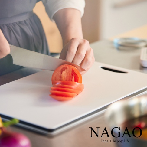 白色雙面抗菌耐熱砧板，搭配切片番茄，上方可見手持菜刀的畫面，砧板邊緣有黑色條紋，右下角有品牌標誌。