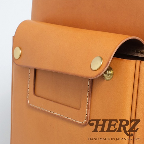 職人手作皮革小學生書包，採用淺棕色皮革，配有兩個金色金屬釦和一個方形標牌，邊緣有縫線裝飾。