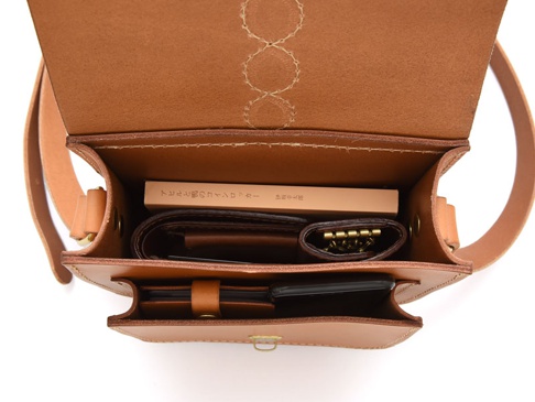 職人手作皮革十字繡紋單肩包，採用原色皮革製作，包包內可見皮夾、鑰匙包和手機，包蓋內側有十字繡紋飾。