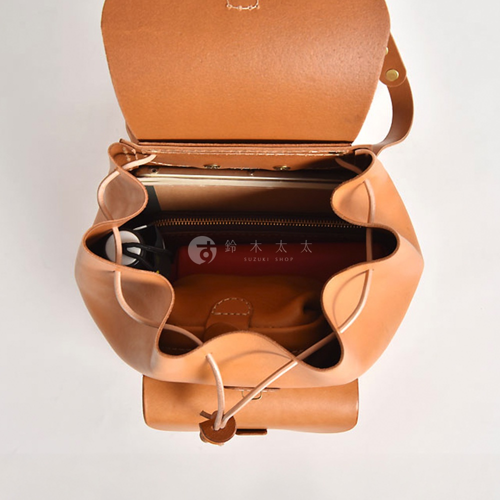 職人手作皮革經典後背包 R-4，此款為淺棕色調，採用天然皮革材質，包身呈現圓潤造型，開口處以抽繩及磁扣設計，內部可見放置的書本、手機及其他小物，展現其收納空間。