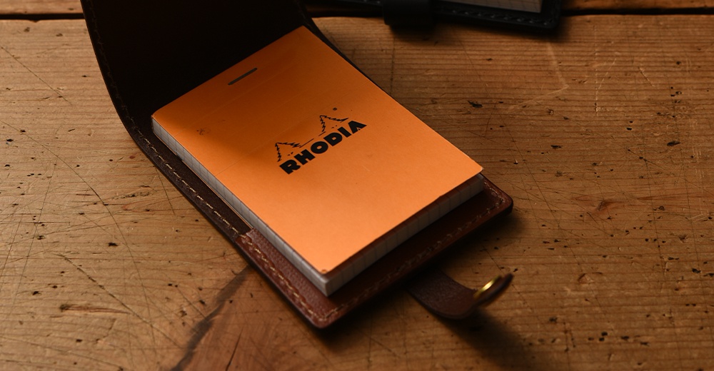 深棕色皮革便利貼收納套，內含一本橘色封面、印有黑色「RHODIA」品牌標誌的筆記本。