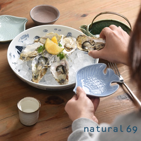 白色陶瓷大圓深碗，碗內飾有藍色魚紋，盛有鋪滿冰塊的新鮮生蠔，點綴著檸檬片與香草。一旁有淺藍色魚造型醬料碟，以及一個白色小陶杯。