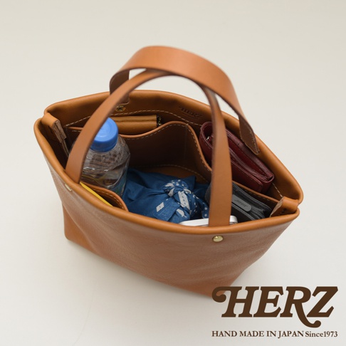 職人手作咖啡色皮革極簡托特包，包內有水瓶、藍色布包、紅色皮夾和灰色手機，展現簡約實用風格。