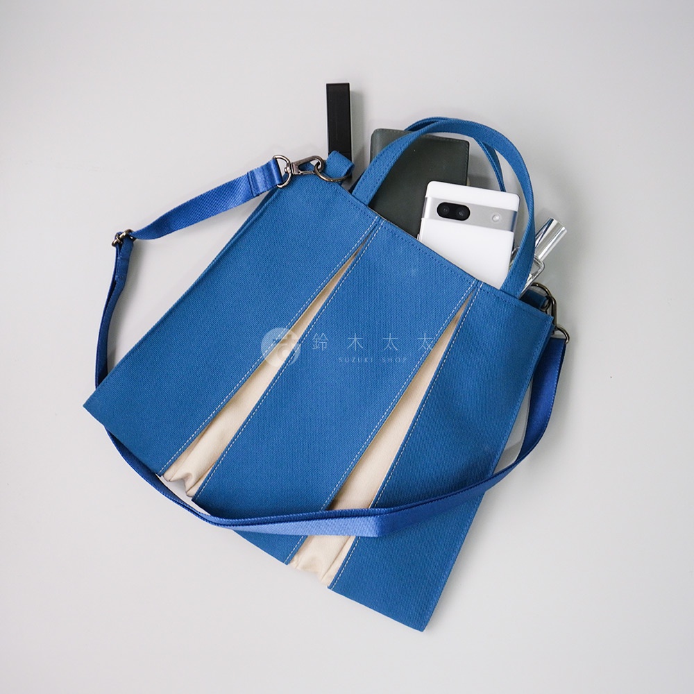 藍色棉帆布扇形托特包，配有米白色內襯，可拆卸肩帶，包內可見手機、筆和卡片夾。