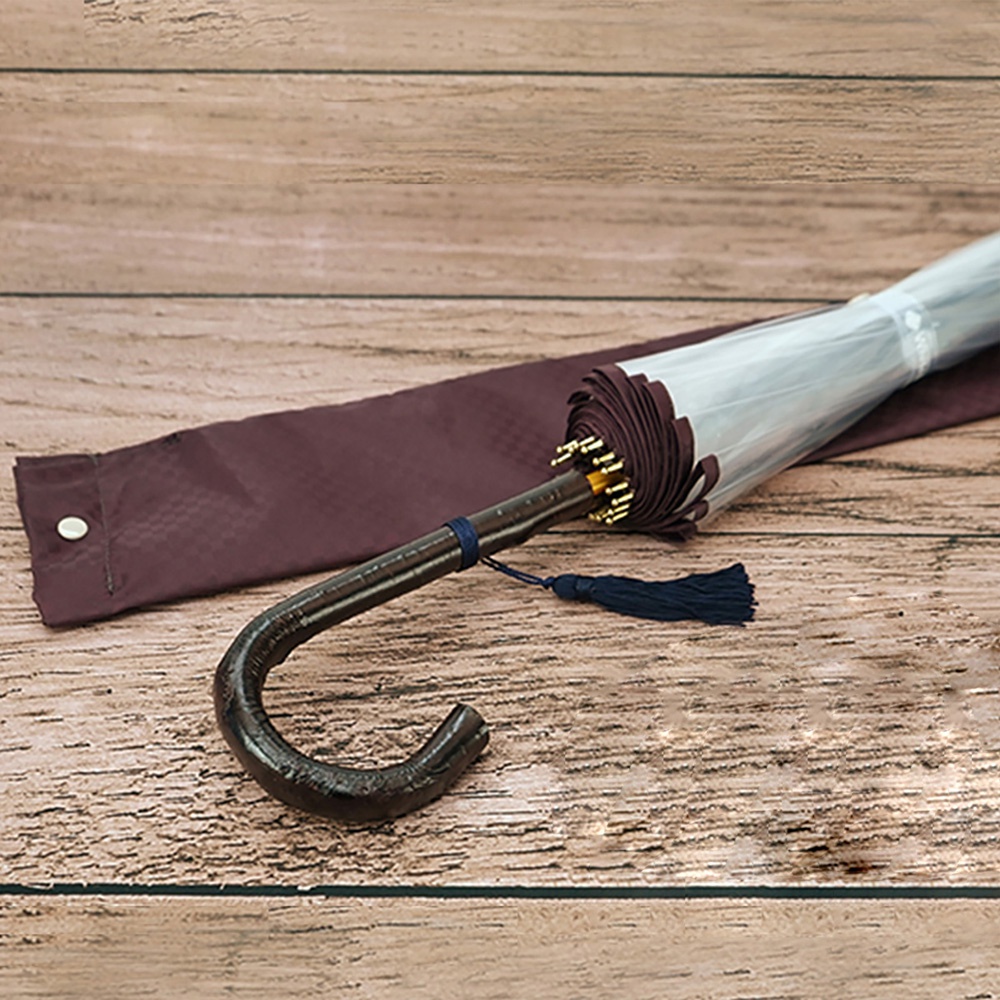 職人高級樺木透明傘，傘骨為深棕色彎曲樺木材質，傘面為透明材質，搭配深藍色流蘇裝飾。傘套為深紅色格紋布料。