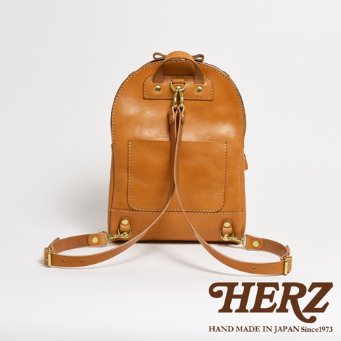 職人手作皮革大開口圓角後背包，顏色為溫暖的焦糖棕色。包款採用天然皮革材質，呈現自然紋理。前方有兩個方形貼袋，背包頂部有提把與可調節的肩帶，皆以金屬扣環固定，展現細膩的職人工藝。