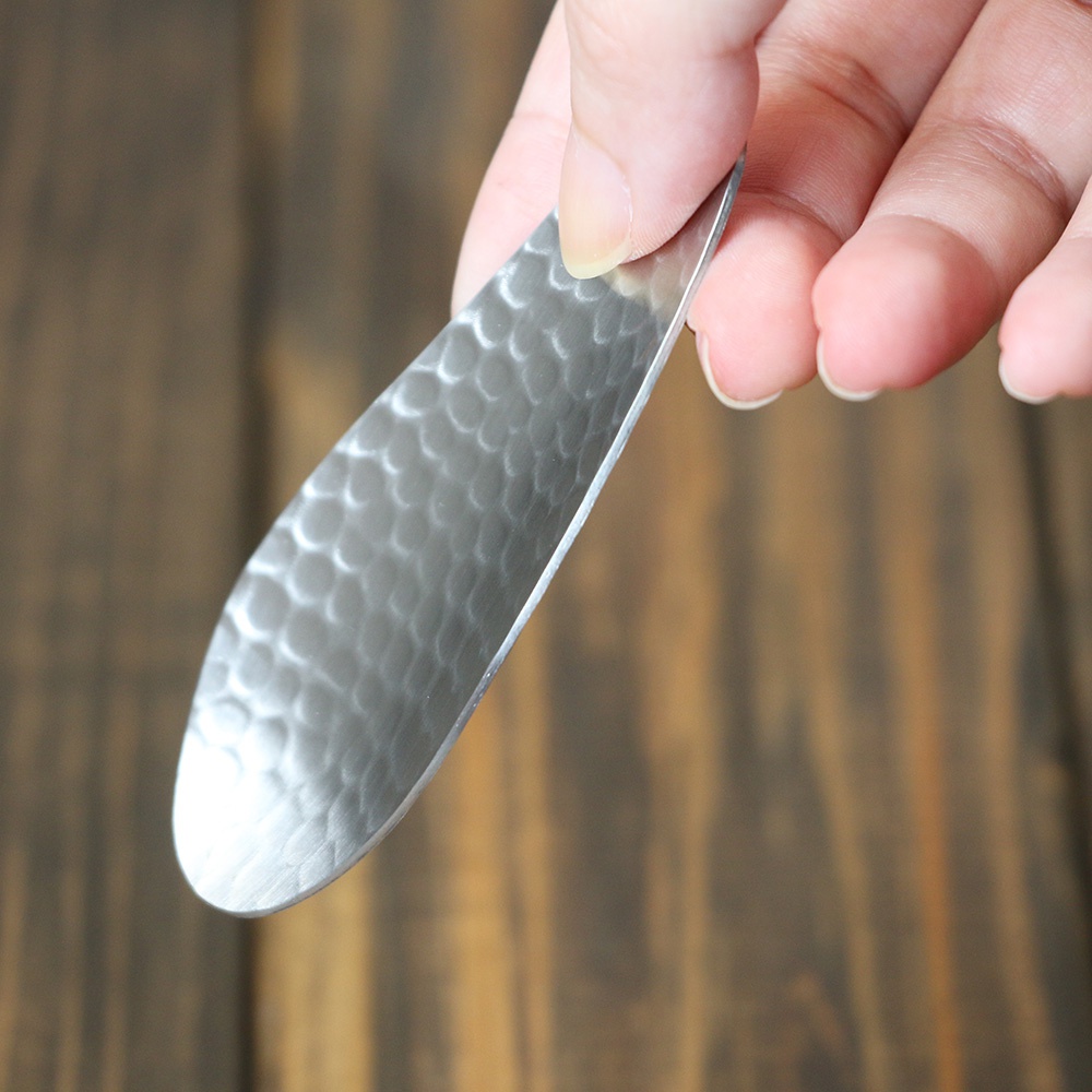 銀色不鏽鋼茶勺，勺面帶有蜂窩狀紋理，由手指握持，背景為木紋桌面。