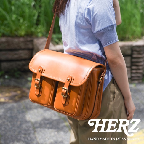 日本HERZ經典雙扣郵差包，以精湛職人手作工藝，打造出質感皮革的雙口袋設計，並印有品牌標誌與「HAND MADE IN JAPAN Since 1973」字樣。