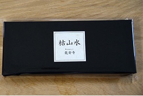 黑色長方形盒裝，表面印有白色方形標籤，標籤上方為「枯山水」字樣，下方為「龍安寺」日文及英文。盒裝置於木紋桌面。