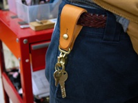 淺棕色皮革製的直立型斜背包，透過銅製釦件固定於深藍色長褲的腰帶環上，並懸掛有兩支鑰匙。