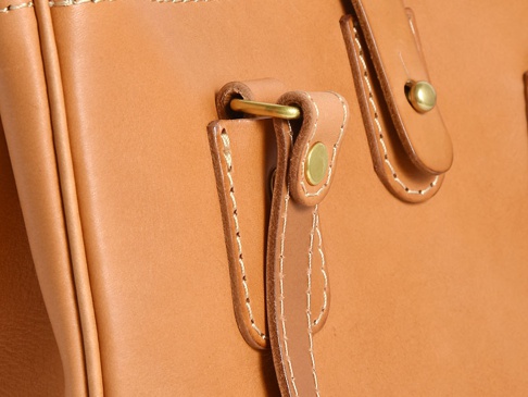 職人手作皮革原子釦托特包，採用天然皮革製成，呈現溫潤的焦糖棕色，縫線細緻，搭配黃銅色金屬釦件，展現簡約質感。