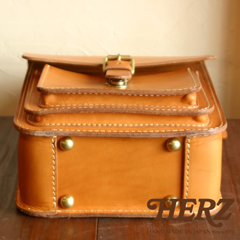 職人手作皮革單肩兩用小方包，採用鞣製皮革材質，呈現經典棕褐色，包身結構層次分明，配有金屬釦環和铆釘裝飾，整體風格簡約復古。