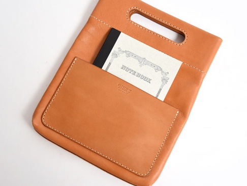 咖啡色皮革直式收納袋，正面有外層口袋，內含一本黑色封面的 NOTEBOOK，袋口上方有提把，皮革表面有縫線裝飾，品牌 logo 壓印在正面口袋上。
