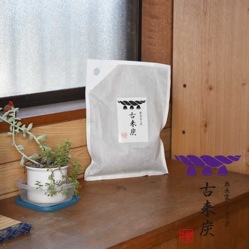 白色包裝的古來炭大型調濕木炭，包裝上印有黑色圖案與「古來炭」字樣。旁邊有綠色植物盆栽，背景為木質紋理。