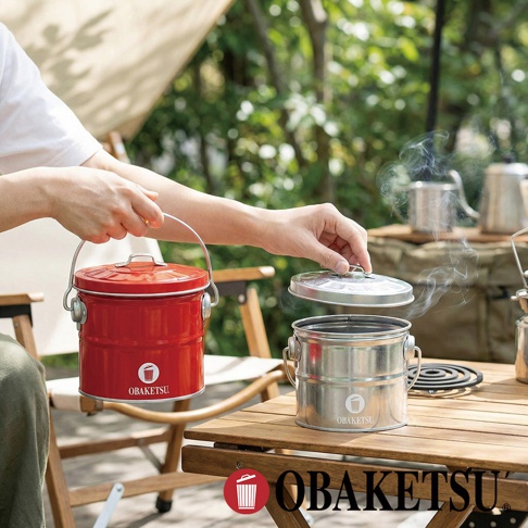 **判斷步驟** 1. 圖片中可讀文字總字數：OBAKETSU（5個字） 2. 由於字數少於50字，選擇模式B。 **輸出** OBAKETSU 品牌的紅色和銀色手工製蚊香桶，放在戶外的木桌上。