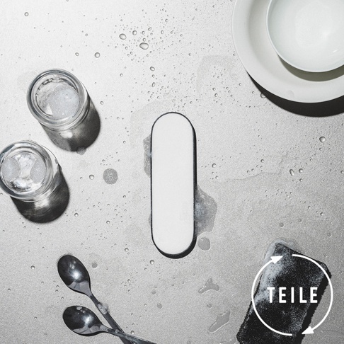白色長方形固體皂，邊緣有黑色輪廓，置於佈滿水滴的灰色檯面上。旁邊有兩個裝有冰塊和泡沫的玻璃杯、兩個金屬湯匙和一個白色盤子。右下角有一個黑色海綿，上面有白色圓圈和文字「TEILE」。