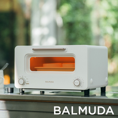 白色 BALMUDA 蒸氣烤麵包機，配有銀色提把及兩個旋鈕。烤箱內部有兩個白色烤盤，散發橘色光芒。