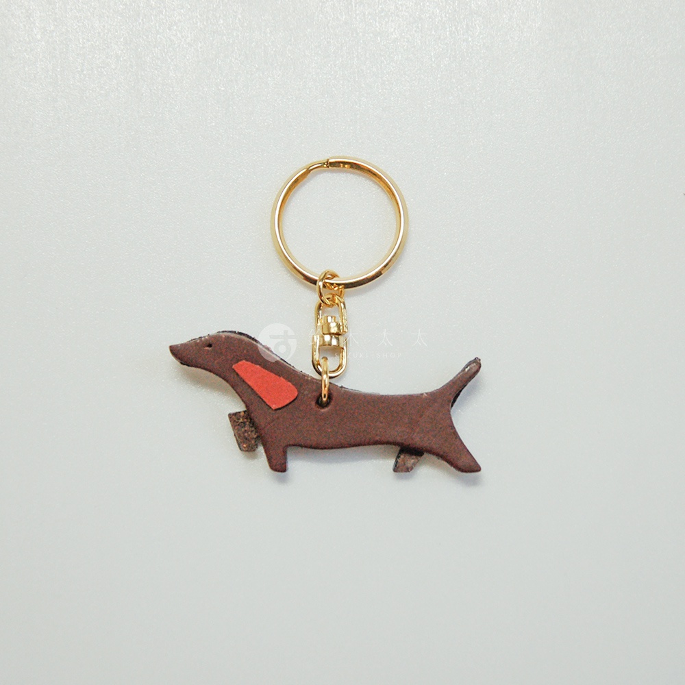 職人手作皮革狗狗造型鑰匙圈，深棕色皮革製成，身體綴有兩塊橘紅色皮革，搭配金色金屬鑰匙圈與掛鉤。