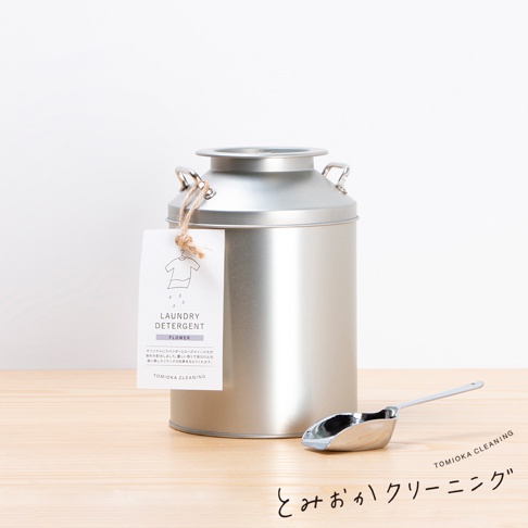 銀色金屬製成的牛奶罐狀洗衣粉收納罐，罐身帶有兩個提把，並掛有印有「LAUNDRY DETERGENT FLOWER」字樣的白色標籤。一旁擺放著一個銀色金屬製的洗衣粉勺。