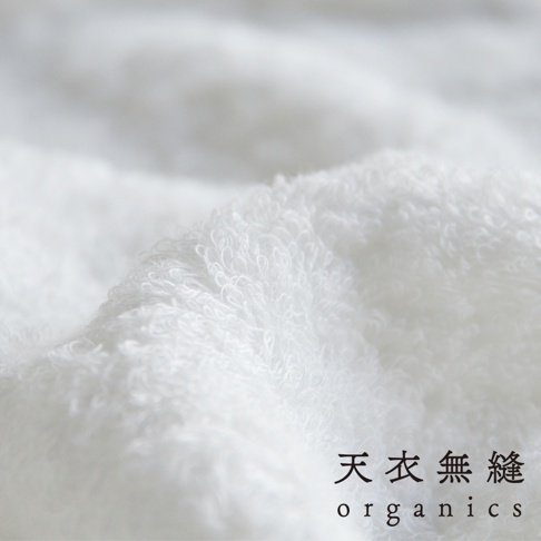 近距離特寫白色蓬鬆柔軟的匹馬棉毛巾，可見細緻的織紋和羽翼般的紋理，底部右側有「天衣無縫 organics」字樣。