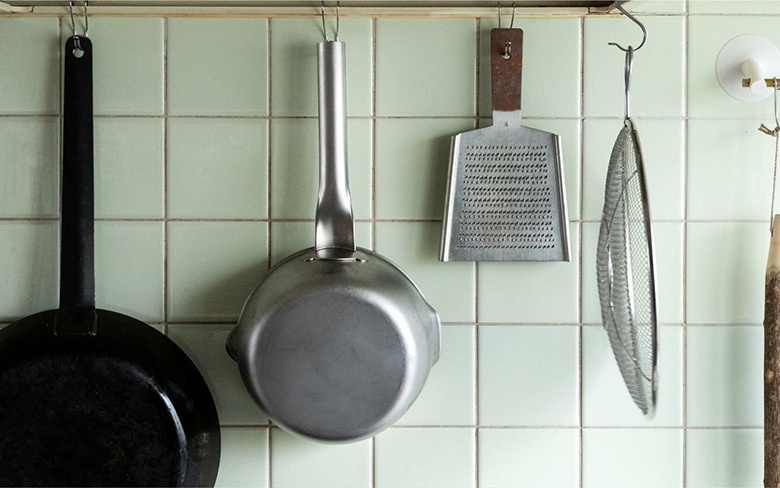 三層不鏽鋼雪平鍋，鍋身呈銀灰色，帶有長柄，懸掛在淺綠色磁磚牆面上。