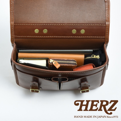 棕色皮革製作的單肩包，內部裝有筆記本、錢包和卡片夾。包包採用紮實的縫線和金屬扣件，設計簡約實用，適合商務或休閒場合。