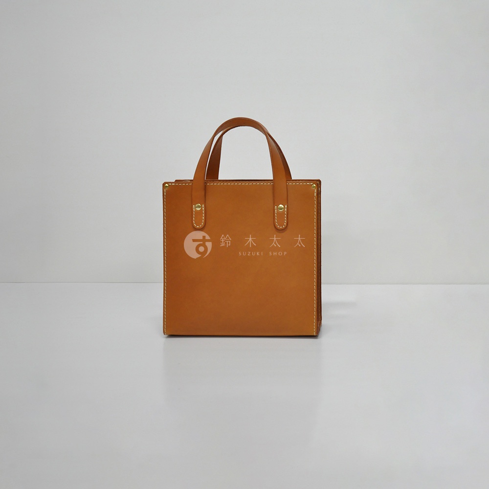 職人手作皮革直式托特包 T-44 XS，採用鞣製皮革製成，呈現經典的棕褐色調。包款設計簡潔，附有兩條手提帶，整體以精緻的縫線收邊，正面中央印有品牌標誌。
