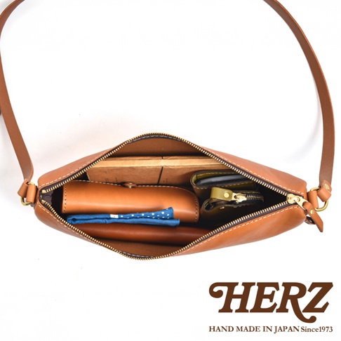 日本 HERZ 職人手作皮革半圓馬鞍包，展示其內部收納，包含皮夾、小錢包、藍色點點手帕及一本木質感的書籍。