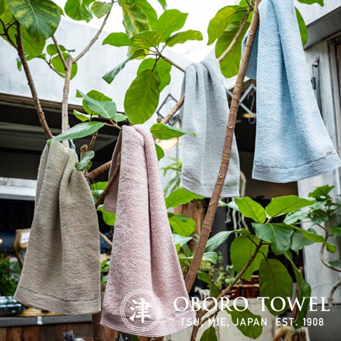 四條不同顏色的毛巾，分別為米色、粉紅色、淺灰色和淺藍色，懸掛在樹枝上，毛巾材質柔軟，帶有細緻的條紋。