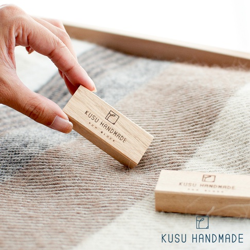 一塊淺棕色、帶有「KUSU HANDMADE ECO BLOCK」蝕刻字樣的木條，由手指拿起，置於淺色與深棕色格紋織物上。另一塊相同的木條放置於織物上。
