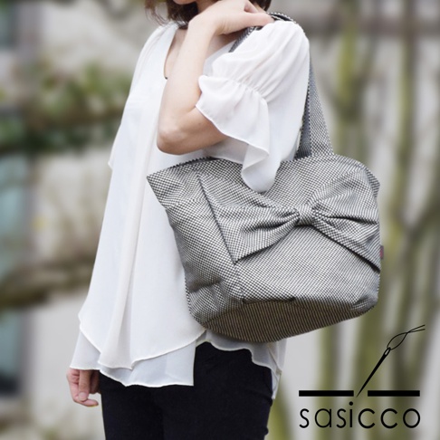 三河木綿刺子織托特包，黑色格紋布料，正面設計有大型蝴蝶結裝飾，搭配白色雪紡上衣與黑色長褲，由一位女士肩背。