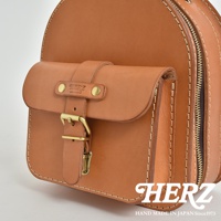 職人手作皮革迷你後背包，採用天然皮革製成，呈現溫潤的棕褐色。包身設計簡潔，配有翻蓋式前袋，以銅釦及皮帶扣固定，側邊設有拉鍊，展現細膩的手工縫線與高品質的五金配件。