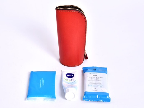紅色皮革直立式筆袋，配有黑色拉鍊，下方擺放著藍色包裝的濕紙巾、白色軟管狀護手霜和另一包藍色包裝的紙巾。