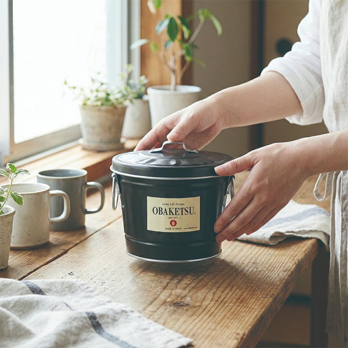 OBAKETSU手工製多功能迷你桶，黑色，附有標籤，標有「OBOKETSU」、「Long Life Design」、「since 1923」、「MADE IN JAPAN」，放置在木桌上，背景有窗戶和綠色植物