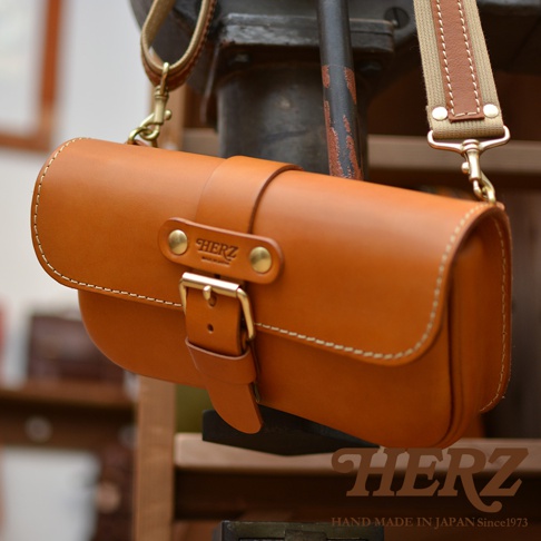 職人手作皮革單扣經典斜背包，採用鞣製皮革製成，顏色為溫暖的棕褐色，配有金色金屬扣環和可調節的帆布與皮革拼接背帶。