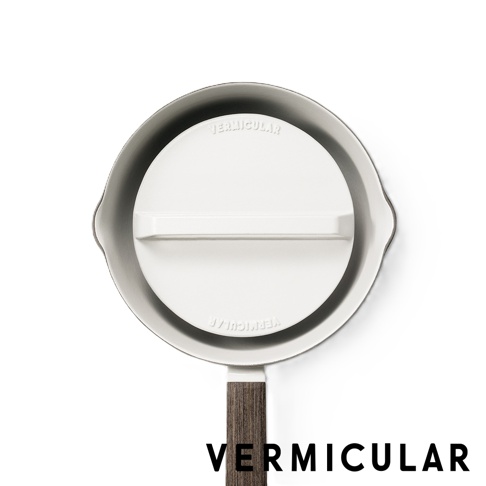 這是一張俯視角度的照片，展示了 Vermicular 品牌的白色琺瑯鑄鐵落蓋，其邊緣略顯灰色，中央配有白色橫向手柄。落蓋表面刻有清晰的 Vermicular 品牌字樣，下方則有一個帶有深色木紋質感的平底鍋手柄延伸出來，整體的設計簡約且具有現代感。