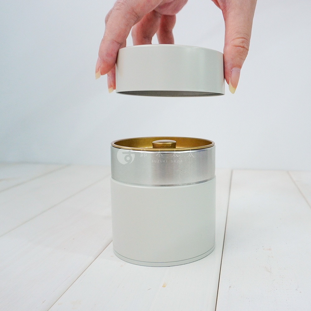 簡約錫製茶葉罐，主體為淺灰色，頂部和中部有銀色飾帶，內緣為金色。罐身光滑，頂蓋由手指拿起，準備蓋上。