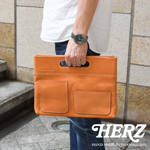 一位穿著白色上衣與藍色牛仔褲的男子，手持一個由日本HERZ品牌職人手作的棕色皮革收納袋，袋身具備兩個外置口袋與精緻的手縫線細節，設計簡約且質感厚實。