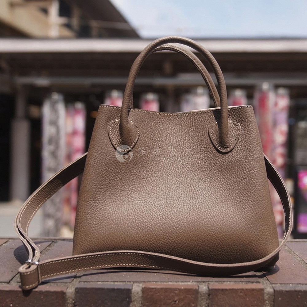 一個棕褐色的皮革兩用手提托特包，具有紋理表面、圓形提把和可拆卸肩帶。