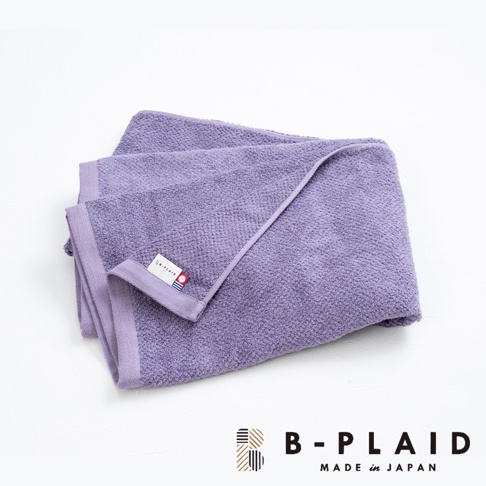 一條淺紫色、織有鱗紋的薄手浴巾，材質觸感柔軟，邊緣有同色系包邊，角落處縫有品牌標籤。