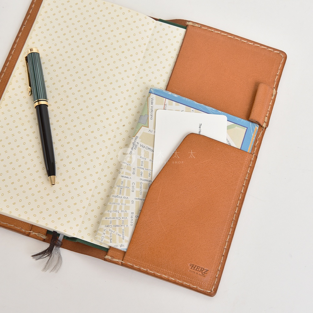 職人手作皮革旅行記事本書套，展示了打開的內頁，內頁有地圖紋飾。書套採用棕色皮革製作，內含多個卡片夾層，並放置有卡片和地圖。一支黑綠相間的筆放在左側內頁上。