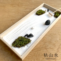 淺木色長方形盆中，舖有白色細沙，沙上耙出細緻紋路。盆內散落幾叢翠綠苔蘚、光滑的黑白卵石，以及一把迷你木耙，構成一幅日式枯山水景觀。