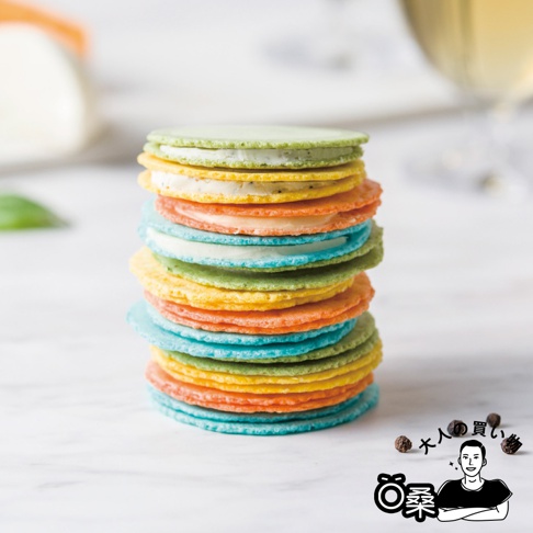 一疊五顏六色的馬卡龍造型仙貝堆疊在白色大理石桌面上，餅體呈現綠色、黃色、橘色與藍色，層層夾著不同風味的起司內餡，右下方繪有黑白線條的卡通人物與大人買物字樣，背景模糊處隱約可見玻璃杯。