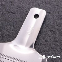 銀色不鏽鋼食材磨泥器，帶有掛孔的長形手柄，手柄上壓印有品牌標誌和「STAINLESS STEEL 5號」字樣。磨泥器部分是平坦的，佈滿鋒利的齒狀磨孔。