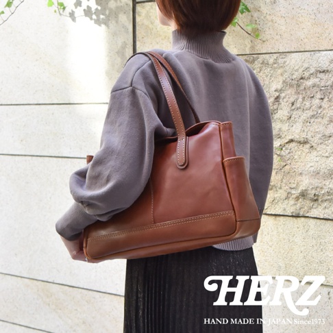 一位女性背著日本HERZ品牌職人手作的棕色皮革肩背托特包，該包款呈現簡約俐落的皮革質感，側面配有細緻的邊袋設計，整體風格優雅且具備手工製作的細節美感。