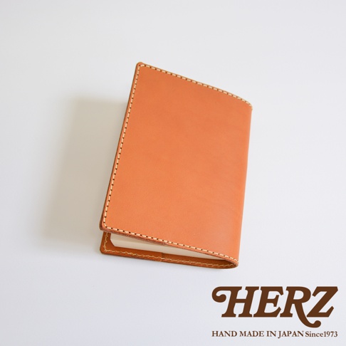 一個淺棕色的日本HERZ職人手作皮革書套，採用厚實皮革製成，表面呈現自然的皮革紋理與溫潤色澤，邊緣以細緻的縫線工藝固定，整體呈現簡約優雅的質感，右下角印有HERZ品牌標誌。