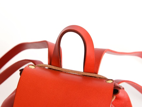 紅色皮革迷你後背包的頂部特寫，展示了手提帶、肩帶連接處的黃銅鉚釘和皮革細節。